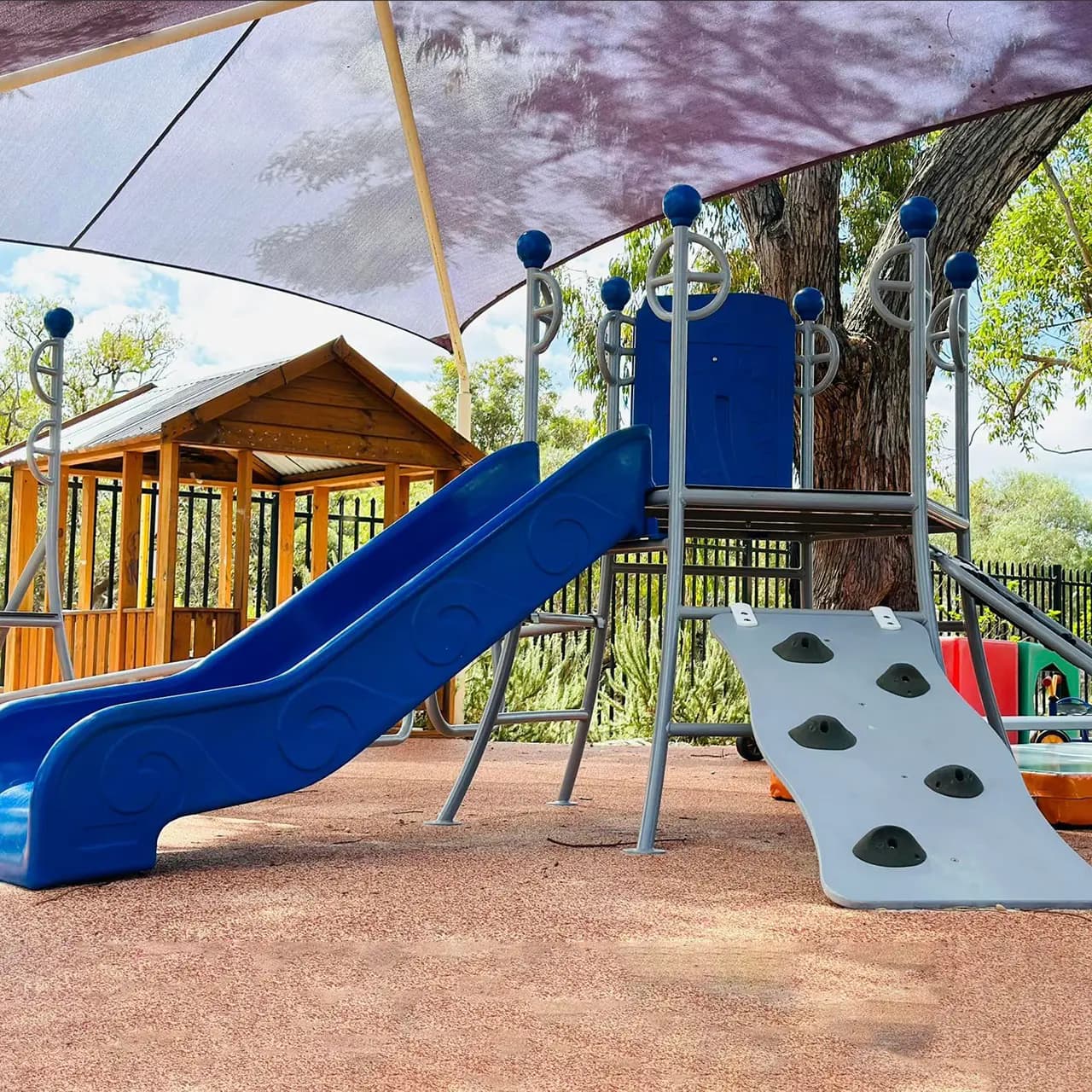 Outdoor play equipment with a blue slide and climbing ramp under a shade sail, showcasing a safe and engaging play environment.